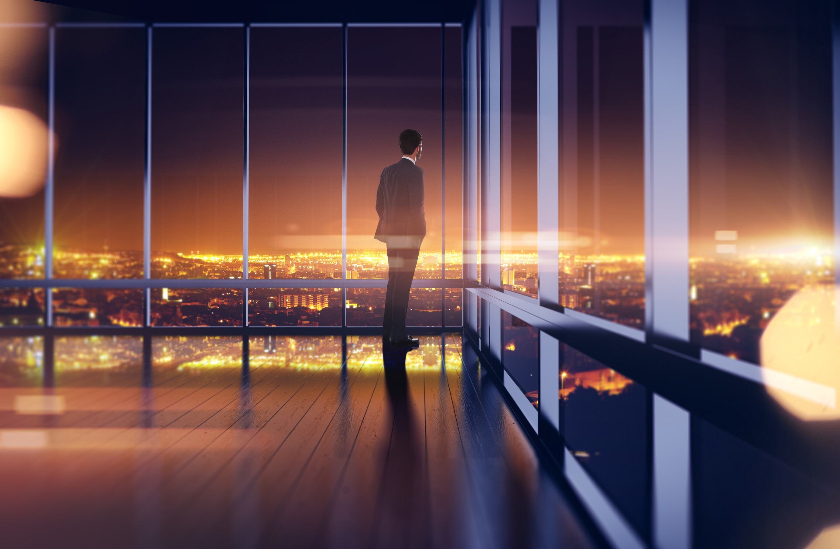 Silhouette of a businessman standing by floor-to-ceiling windows, looking out at a brightly lit city skyline at night.