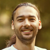 Headshot of a smiling man with long brown hair tied back, wearing a white shirt, photographed outdoors with a blurred natural background.